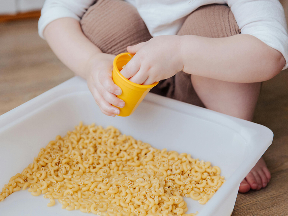 toddler playing with dry pasta in sensory bin 