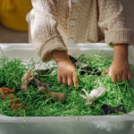 A little girl playing with farm animals in sensory bin with green shredded paper. Educational game, learning through play. Montessori material for toddlers. Sensory play activities