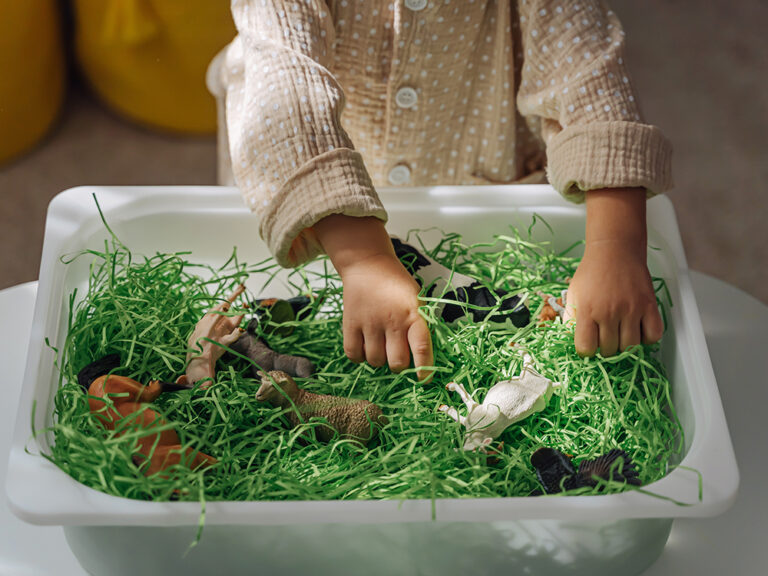 A little girl playing with farm animals in sensory bin with green shredded paper. Educational game, learning through play. Montessori material for toddlers. Sensory play activities